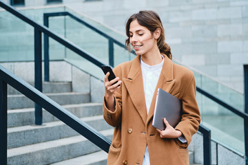 Young casual businesswoman in coat with laptop happily using cellphone outdoor
