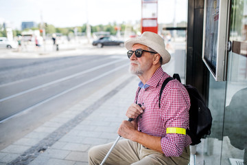 Senior blind man with white cane waiting for public transport in city.