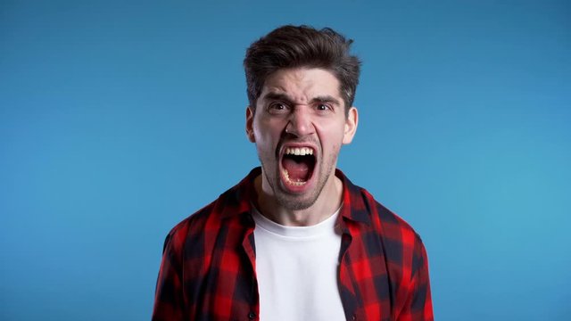 Young european stressed man in red shirt shouting isolated over blue background. Slow motion. Stressed and depressed guy loudly screaming to camera.