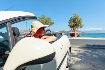 Mature woman with hat and sunglasses driving her convertible top automobile on bright sunny day near ocean.