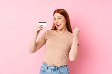 Young redhead woman over isolated pink background holding a credit card