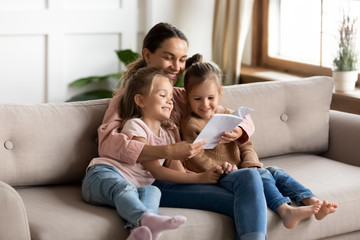 Happy young mother read book embrace little children daughters