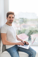 Young man working on laptop and sitting on windowsill