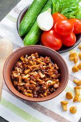 Fried chanterelles with onion in rustic bowl and plate with fresh vegetables for salad on background