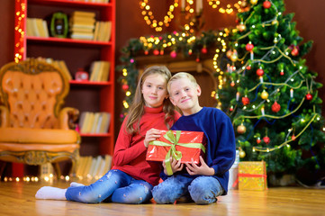 Obraz na płótnie Canvas Sister and brother are sitting on the floor in the room with a gift in their hands. Against the background of a festive tree. Children give presents. Christmas and New Year