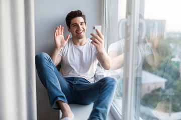 Young handsome man sitting on the windowsill make video call on the phone