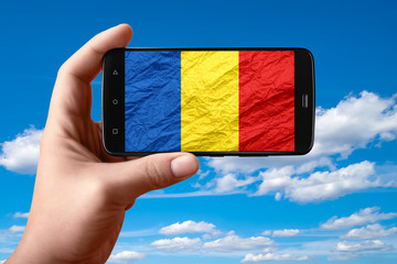 Romania flag on the phone screen. Smartphone in hand shows a flag on a background of the sky with...
