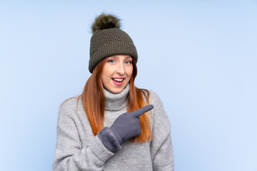 Young redhead Russian woman with winter hat over isolated blue background pointing finger to the side