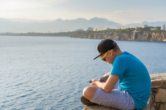 Closeup profile view of handsome white kid using modern mobile smartphone sitting in beautiful scenic summer sunny landscape of Turkey, Antalya city. Horizontal color photography.