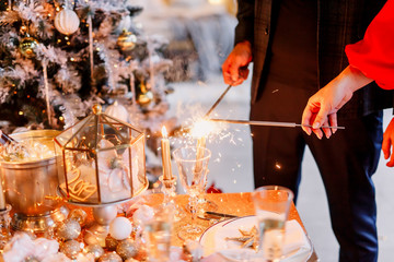 New Year 2020. New Year mood. Table with a gold tablecloth, decorated with candles and tableware for celebration. Festive still life by candlelight. A young couple lights sparklers. Close-up.