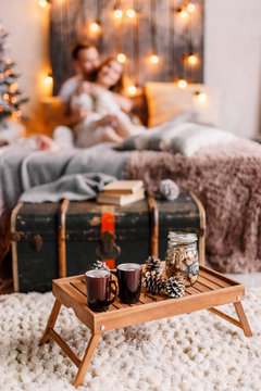 Table with cup on the plaide, marshmallow and Christmas cookies, cozy knitted blanket. New Year 2020. Composition with a cup tray & a mug. Husband and wife in the background are hugging on the bed.