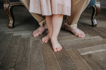 Legs of unrecognizable little girl and dad barefoot standing on the wooden floor.