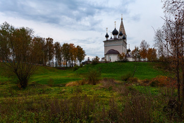 An Orthodox white-stone church with black domes and golden crosses stands on top of a hill against a background of rain sky and autumn trees