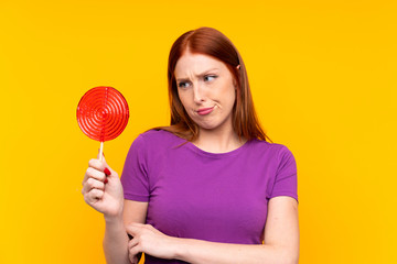 Young redhead woman holding a lollipop over yellow background with sad expression