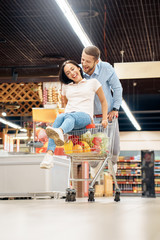 Daily Shopping. Couple in the supermarket together man walking with cart while woman sitting inside having fun laughing cheerful