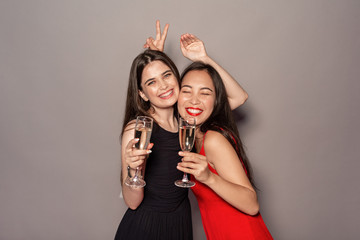 Freestyle. Young girls in dresses standing isolated on gray with glasses of champagne gesturing playful laughing cheerful