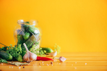 Pickling cucumbers in cans. Ingredients for marinated gherkins, glass jar, garlic, dill, salt, pepper on yellow background. Copy space. Concept of vegetables preservation.