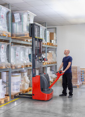Warehouseman in storehouse moving pallet with electric stacker. Worker in warehouse stacks pallet in rack system.Caucasian labourer working with small electric forklift. Man with high lift pallet jack - obrazy, fototapety, plakaty