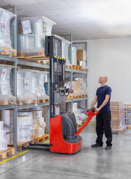Warehouseman in storehouse moving pallet with electric stacker. Worker in warehouse stacks pallet in rack system.Caucasian labourer working with small electric forklift. Man with high lift pallet jack
