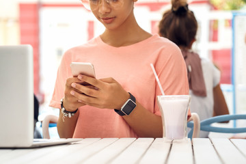 In the Cafe. Young girl in glasses sitting at table with milkshake using laptop browsing smartphone close-up