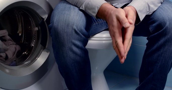 part of the bathroom. a man in jeans sits on a closed toilet next to a washing machine. he rubs his palms together, interlacing his fingers