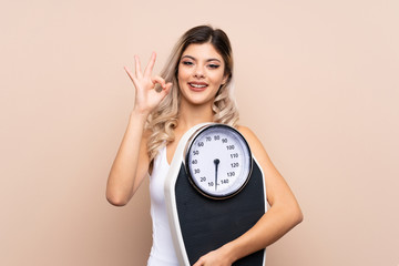 Teenager girl with weighing machine over isolated background holding a weighing machine and doing OK sign - obrazy, fototapety, plakaty
