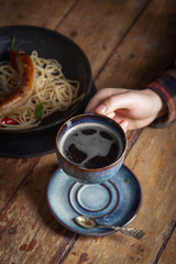 Hearty rustic bachelor breakfast Italian pasta spaghetti, bitten fried meat sausage, red chili pepper in black frying pan, fork. Still life. Lonely man lunch, black americano coffee cup in hand