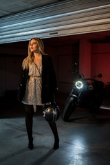 Fototapeta na wymiar Attractive blonde woman posing for fashion photo shoot in a garage with elegant dress and a motorcycle behind