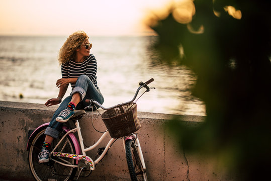 Adult caucasian woman enjoying outdor leisure activity relaxing after a ride on a vintage bike, sitting on a wall looking at the ocean -healthy active lifestyle people