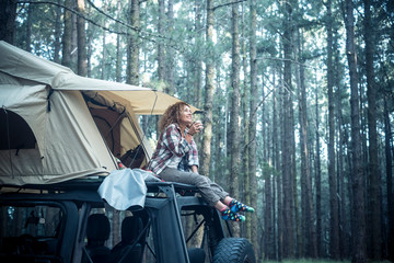 Happy free beautiful people caucasian woman sit down on the roof of the car with tent mounted on it for free travel camping lifestyle - wanderlust vehicle to enjoy the nature and vacation in forest