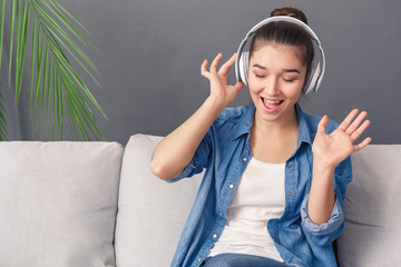Hobby Concept. Young woman in headphones sitting on sofa studio isolated on grey listening song singing along smiling joyful