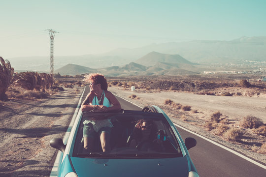 Couple of happy friends adult caucasian women drive and travel on a blue convertible open car together in friendship - holiday vacation concept with long asphalt road and landscape in background