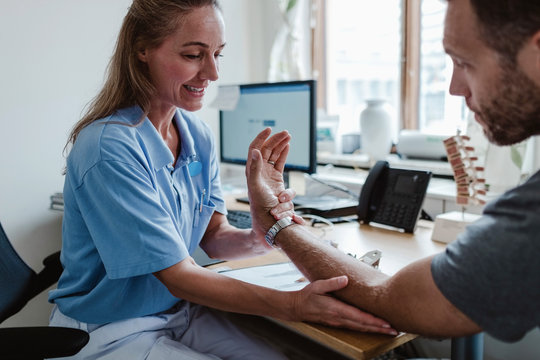 Smiling doctor examining male patient's hand while sitting in medical examination room at clinic