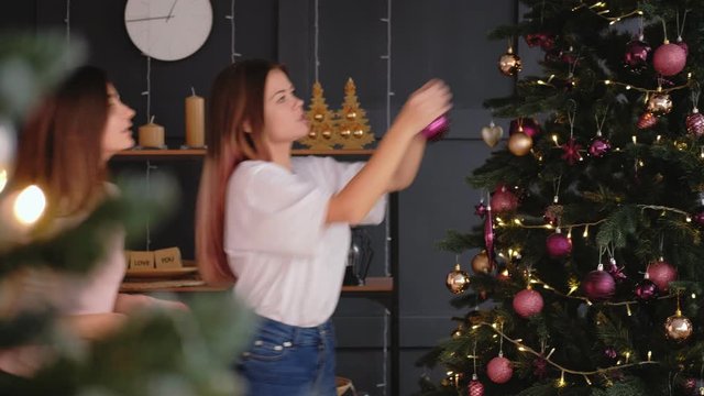 Winter holidays preparations. Christmas Eve. Two happy teen girls decorating fir tree with baubles.