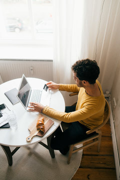 High angle view of man with credit card using laptop while sitting at table in living room