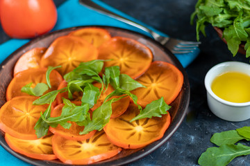 Persimmon Salad. Thinly sliced fruit slices lie on a brown plate, sprinkled with arugula leaves. Vegetarian food. Copy space. 