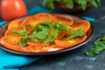 Persimmon Salad. Thinly sliced fruit slices lie on a brown plate, sprinkled with arugula leaves. Vegetarian food. Copy space. 