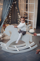 Fototapeta na wymiar Stock photo of adorable little kid enjoying time sitting on a white wooden horse in room decorated for Christmas.