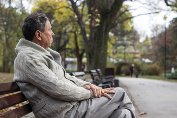 Portrait of a pensive senior man sitting on the bench, in the public park, outdoors. Old man relaxing outdoors and looking away. Portrait of elderly man enjoying retirement