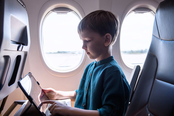 Cute six years old boy, playing on tablen in aircraft on boar, traveling on vacation with parents and siblings
