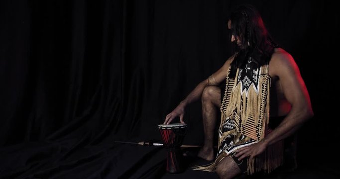 Native american with long hair is sitting on the floor next to a spear and drum