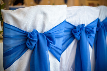 Decorated chairs with black fabric and big blue satin bows. Silk bow tied on back of chair in party at restaurant. Dark cloth cover chairs standing in a row