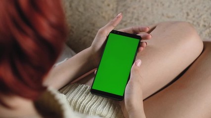 Woman uses green screen online. adult girl is sitting on couch and holding modern phone with green screen. Chromakey on the smartphone. Use the green layout to insert images. Hand and finger gestures.