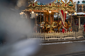 Сarousel horses to traditional Christmas market. Outdoor fair on winter day. View through the frost pine branch. Happy new year. Festive street decor in winter holidays.