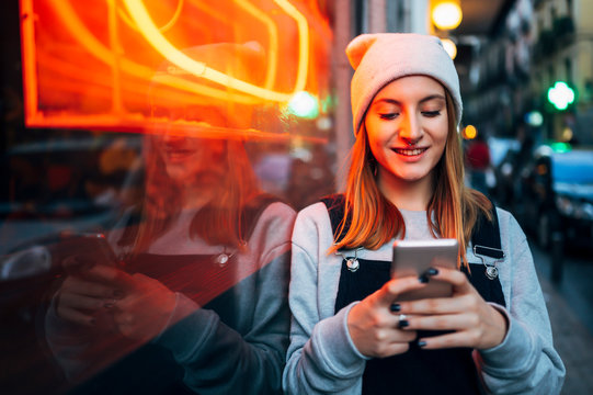 Smiling young woman standing next to neon light using her smartphone at night