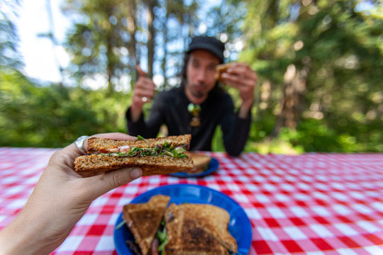 Selective focus of two adult people having a breakfast or lunch at a picnic table eating hot sandwiches while spending time in nature