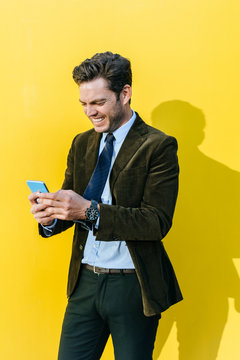 Happy businessman using smartphone in front of yellow wall