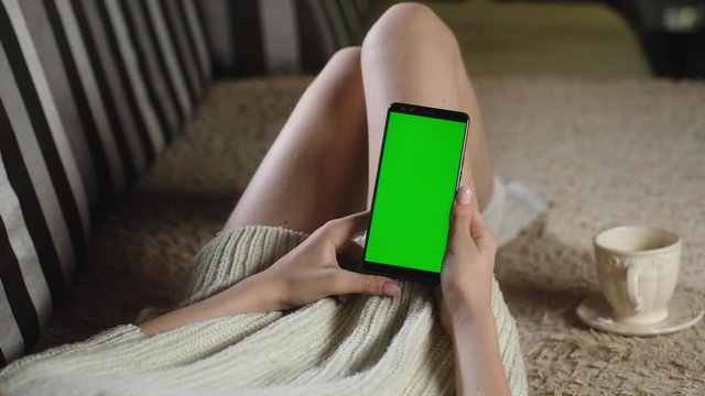 girl uses phone with green screen. chromakey smartphone. woman in sweater sits on couch with Cup of coffee and holds modern smartphone. Use green mockup insert images. swipe photos with your fingers.