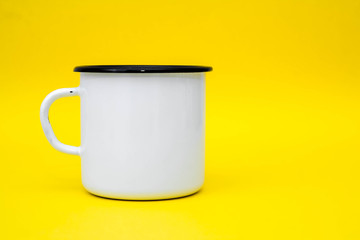 Enamel white metal mug with black line on the edge on yellow background. Mug empty blank for coffee or tea. Space for text