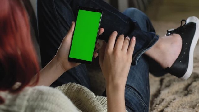 girl gesture flips through photos with her fingers using her green screen phone. smartphone chromakey. woman is resting on couch and holding smartphone. Use the green mockup to insert photo or image.